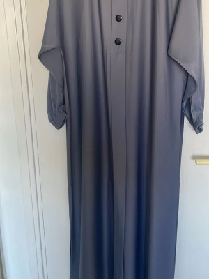 Lång klänning med knappar - En stilren långklänning i grått med vida ärmar och två svarta knappar framtill. Klänningen har en lös passform och resår vid ärmsluten för en avslappnad look. Perfekt för dig som gillar minimalistisk stil. Färgen är mellan lite blå och grå, PRIS GÅR ATT DISKUTERAS! 