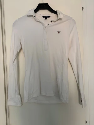 Vit långärmad henley från Gant - Vit långärmad henley från Gant med klassisk krage och knappar vid halsen. Skjortan har en liten broderad logga på bröstet och är tillverkad i mjuk bomull. Perfekt för en avslappnad och stilren look. Små fläckar på magen som knappt syns i verkligheten. 