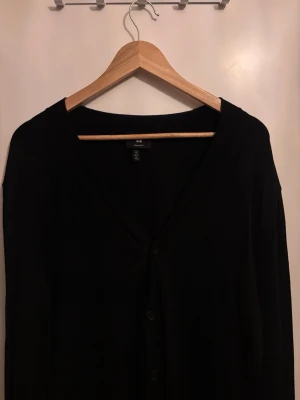 Svart kofta från H&M med knappar - En klassisk svart kofta från H&M med v-ringning och knäppning framtill. Långärmad och i mjukt material, perfekt att slänga på sig över en t-shirt eller skjorta. Enkel och stilren design som funkar till det mesta.