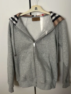 Grå zip hoodie från Burberry - Snygg grå zip hoodie från Burberry med klassiskt rutigt mönster i huvan och broderad logga på bröstet. Tillverkad i mjuk bomullsblandning, har dragsko i huvan och två fickor framtill. Perfekt för en chill och stilren look. Storlek M men passar om due runt 167-178. Kom gärna med prisförslag.