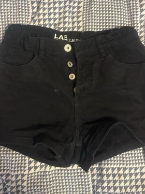 Svarta högmidjade shorts med knappar - Svarta högmidjade shorts från LAB Industries med fyra synliga knappar framtill. Klassisk femficksmodell i bomull, perfekt till sommaren. Stilsäker och enkel design som passar till det mesta.