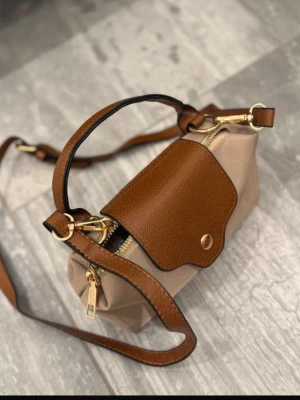 Beige och brun handväska med axelrem - Snygg handväska i beige tyg med bruna detaljer.Väskan har guldfärgade metalldetaljer, justerbar axelrem och ett lock med tryckknapp. Perfekt storlek för det viktigaste och en stilren design som passar till många outfits.