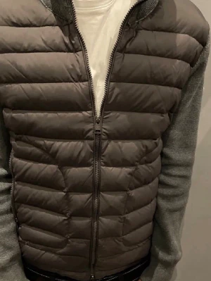 Moncler grå dunväst med stickade ärmar - Snygg grå dunväst från Moncler med quiltad front och rygg samt stickade ärmar i mörkgrått. Västen har dragkedja framtill och Moncler-logga på ärmen. Perfekt för lager-på-lager och ger en modern look.