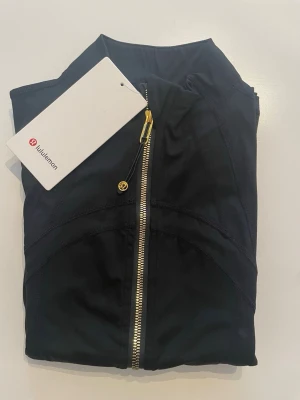 Svart träningsjacka från Lululemon - Snygg svart träningsjacka från Lululemon med guldfärgad dragkedja och hög krage. Figurnära passform och långa ärmar. Tillverkad i ett stretchigt funktionsmaterial som andas, perfekt för träning eller uppvärmning.