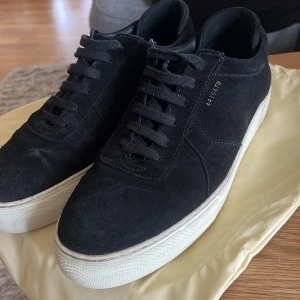 Svarta sneakers från Axel Arigato - Snygga svarta sneakers från Axel Arigato i mocka med vit platt sula. Klassisk och stilren design med snörning och diskret guldfärgad logga på sidan. Perfekta för dig som gillar minimalistisk stil och vill ha ett par riktigt schyssta skor till vardags.