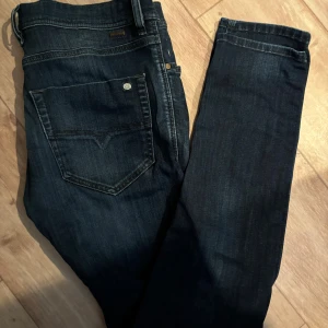 Mörkblå jeans från Diesel - Säljer ett par mörkblå jeans från Diesel med klassisk femficksdesign och raka ben. Jeansen har snygga slitningar framtill och kontrastsömmar. Perfekta för en avslappnad och trendig look.