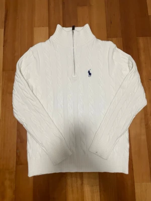Vit stickad tröja Polo Ralph Lauren - Kabelstickad vit tröja från Polo Ralph Lauren med halv dragkedja och hög krage. Klassisk blå logga broderad på bröstet och ribbade muddar vid ärmslut och nederkant. Perfekt för dig som gillar stilren och tidlös design.