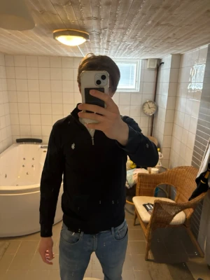 Svart halvzip från Ralph Lauren - Snygg svart tröja från Ralph Lauren med halvzip och den klassiska vita loggan broderad på bröstet. Tröjan har lång ärm och en smal passform som passar perfekt till jeans eller chinos. Enkel och stilren design som funkar året runt.