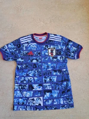 Blå Japan Adidas anime t-shirt - Unik blå t-shirt från Adidas med Japans landslagsemblem och animeinspirerat mönster över hela tröjan. Röd kant vid hals och ärmslut, samt vita ränder på axlarna. Perfekt för dig som älskar både fotboll och anime.