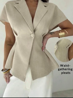 Beige kavaj med markerad midja - Stilren beige kavaj med korta ärmar och markerad midja tack vare veckade detaljer. Kavajen har en enkel knapp framtill och klassisk krage. Materialet ser ut att vara en lätt, vävd textil som ger en elegant och modern look.