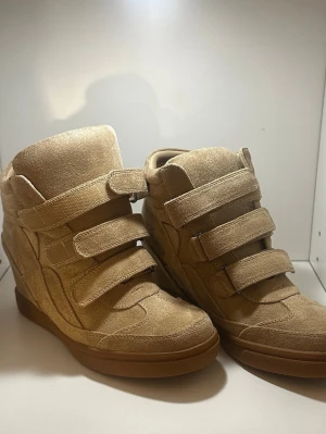 Beige sneakers med kardborreband - Snygga beige sneakers i mocka-look med tre breda kardborreband och bruna gummisulor. Skorna har en hög siluett och vadderad plös för extra komfort. Perfekta för dig som vill ha en clean och trendig stil.