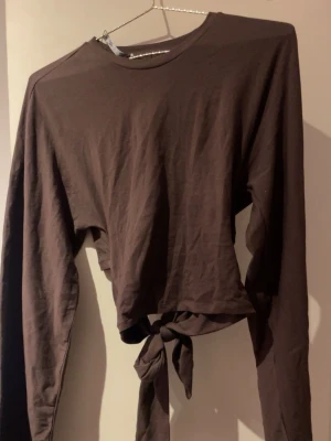 Brun croppad topp med knyt - Snygg brun croppad topp med långa ärmar och knyt i midjan. Tillverkad i mjukt bomullstyg och har en enkel, stilren design utan mönster eller logga. Köpt i lager157 men helt oanvänd utan prislapp.🩷