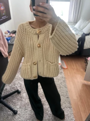Chunky beige stickad kofta - Säljer en chunky stickad kofta i beige med stora guldknappar och två fickor framtill. Koftan har en grovstickad struktur, rund halsringning och långa ärmar. Perfekt för lager-på-lager och en mysig look.