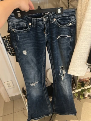 Blå slitna bootcut jeans - Säljer ett par blå bootcut jeans med slitningar och trasiga detaljer på benen. Jeansen har klassiska fem fickor och kontrastsömmar. Perfekt för dig som gillar en cool och avslappnad look.