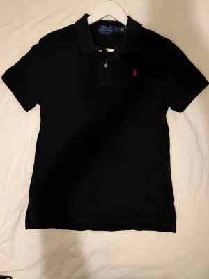 Svart pikétröja från Polo Ralph Lauren - Klassisk svart pikétröja från Polo Ralph Lauren i slim fit-modell. Tröjan har korta ärmar, krage med knappar och den ikoniska röda logotypen broderad på bröstet. Perfekt till jeans eller shorts för en clean och stilren look.