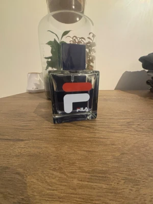 Fila parfym i fyrkantig flaska - Stilren parfym från Fila i en fyrkantig glasflaska med stor blå kork. Flaskan har en modern design med Fila-loggan i vitt och rött på framsidan. Perfekt för dig som gillar sportiga och fräscha dofter. Lite använd men mycket kvar, ny pris ligger på ungefär 229kr.