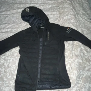 Svart pufferjacka från Sail Racing - Svart pufferjacka från Sail Racing med huva och dragkedja. Jackan har quiltade partier framtill och på ryggen, samt logga på bröstet och ärmen. Långärmad modell med sportig look, perfekt för kyliga dagar. Har haft jackan inte så länge kanske i typ 4-5 månader, kan diskutera pris!