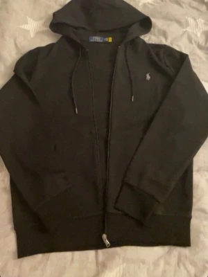 Svart hoodie från Polo Ralph Lauren - Snygg svart hoodie från Polo Ralph Lauren med dragkedja framtill och klassisk huva med dragsnören. Liten broderad logga på bröstet. Tillverkad i mjukt material som känns skönt mot huden. Perfekt för en avslappnad och stilren look.