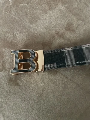 Burberry bälte  - Snyggt bälte med rutigt mönster i svart och grått samt ett stort guldfärgat B-spänne. Perfekt accessoar för att lyfta din outfit med en cool detalj. Bältet har en klassisk design och känns både trendigt och stilrent.