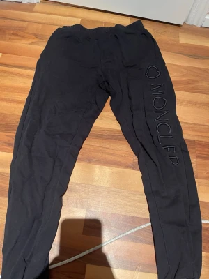 Svarta mjukisbyxor från Moncler - Säljer ett par svarta mjukisbyxor från Moncler med broderad logga och text längs ena benet. Byxorna har resår i midjan och är tillverkade i mjukt material som passar perfekt för en avslappnad stil. Snygg och enkel design med diskreta detaljer. OBS: 2 hål i byxorna inte synliga