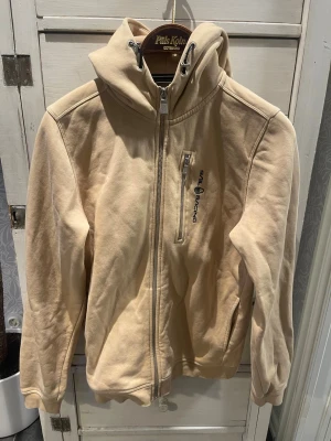 Beige hoodie från Sail Racing - Beige hoodie från Sail Racing med dragkedja framtill, huva med dragsko och en bröstficka med vertikal dragkedja. Logotyp på bröstet och en detalj på huvan. Tillverkad i mjukt bomullsmaterial, perfekt för en avslappnad stil.