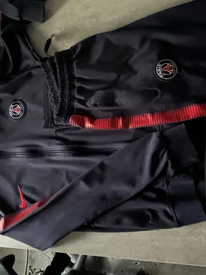 Mörkblå PSG träningsset från Nike - Säljer ett  mörkblå träningsset från Nike x PSG med röda detaljer. Setet består av en zip-jacka och matchande byxor, båda med PSG-logga och Nike swoosh. Materialet är mjukt och stretchigt, perfekt för träning eller chill. Röd rand längs ärmar och ben ger extra stil.