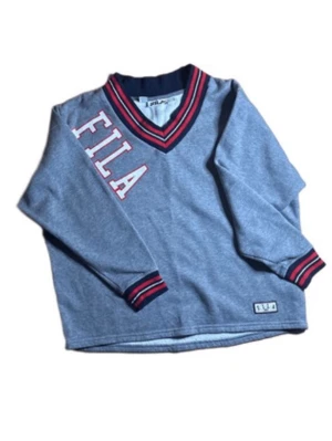 Grå stickad FILA tröja - Snygg grå stickad tröja från FILA med röd och marinblå randig V-ringad krage och muddar. Stor FILA-logga i vitt och rött på bröstet samt en liten logotyp nertill. Långärmad och i mjukt material, perfekt för en sportig och avslappnad look.