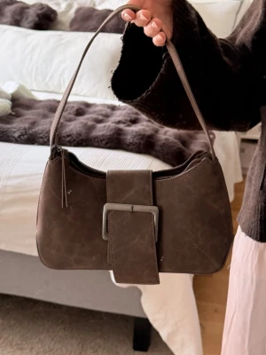 Brun axelremsväska med spänne - Snygg brun handväska i fejkläder med axelrem och stort dekorativt spänne framtill. Väskan har en mjuk, rundad form och stängs med dragkedja. Insidan är fodrad i matchande brunt tyg och har en innerficka med dragkedja. Perfekt accessoar för en trendig look.