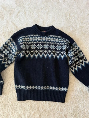 Mörkblå stickad tröja i Ull - Säljer en mörkblå stickad tröja med klassiskt nordiskt mönster i vitt och ljusblått. Tröjan har rund halsringning, långa ärmar och ribbade muddar vid ärmslut och nederkant. Perfekt för kalla dagar och riktigt snygg till jeans.