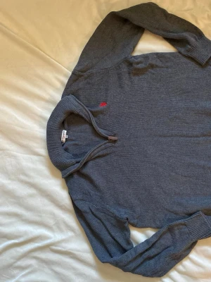 Mörkgrå stickad tröja från Polo assn  - Mörkgrå stickad tröja från Polo assn med halv dragkedja och ribbade muddar. Tröjan har en liten röd broderad logga på bröstet och är tillverkad i mjukt material. Perfekt för lager på lager och en stilren look.