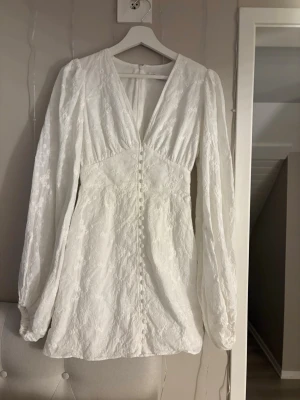 Vit spetsklänning med knappar - Romantisk vit klänning med v-ringning, långa puffärmar och dekorativa knappar framtill. Klänningen har spetsdetaljer och en figurnära passform som ger en elegant siluett. Perfekt för speciella tillfällen eller studentfirande.