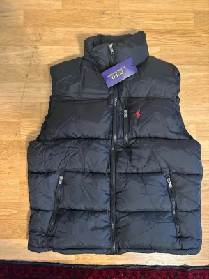 Svart dunväst från Polo Ralph Lauren - Snygg svart dunväst från Polo Ralph Lauren med hög krage och dragkedja framtill. Västen har två framfickor med dragkedja och den klassiska röda Polo-loggan broderad på bröstet. Perfekt för lager på lager och streetwear-vibe.