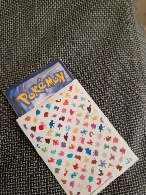 Pokémonkort Stakataka GX Ultra Beast - Säljer ett Pokémonkort med motivet Stakataka GX Ultra Beast. Kortet har metallic-effekt och är tryckt i färgerna svart, röd, grå och silver. Ingår även ett ark med små färgglada Pokémon-klistermärken i olika former och färger. Perfekt för samlare eller Pokémonfans.