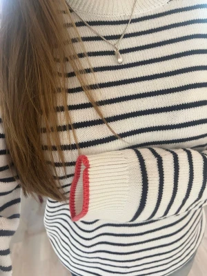 Randig stickad tröja med knappar - Vit stickad tröja med svarta horisontella ränder och svarta knappar på axeln. Ärmsluten har en röd detalj som kontrast. Tröjan har rund halsringning och är långärmad. Perfekt för en stilren och avslappnad look.