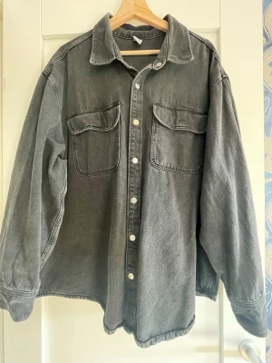 Oversized svart jeansjacka - Snygg oversized svart jeansjacka med två stora bröstfickor och silverfärgade knappar framtill. Klassisk krage och avslappnad passform. Tillverkad i kraftigt denimtyg som ger en cool vintagekänsla. Perfekt att slänga över en hoodie eller t-shirt.