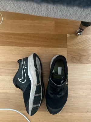 (Barn )Svarta Nike sneakers med vit sula - Snygga svarta Nike sneakers med vit sula och klassisk swoosh på sidan. Skorna har meshmaterial upptill för bra ventilation och kardborreband över snörningen för extra support. Perfekta för en sportig och avslappnad stil.