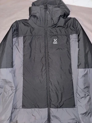 Svart och grå dunjacka från Haglöfs - Snygg svart och grå dunjacka från Haglöfs med huva och hel dragkedja framtill. Jackan har två sidofickor med dragkedja och diskret Haglöfs-logga på bröstet. Perfekt för kalla vinterdagar och tillverkad i slitstarkt material.