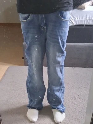 Blå baggy jeans med färgstänk - Säljer ett par ljusblå baggy jeans med vita färgstänk och raka ben. Jeansen har klassisk femficksdesign och är tillverkade i denim. Perfekta för en avslappnad och trendig streetwear-look.