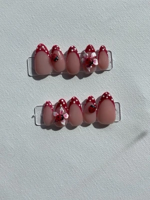 Press-on naglar med nyckelpigor & blommor - Säljer ett set press-on naglar i mandelform med nude bas och röda toppar dekorerade med vita prickar. Några naglar har 3D-blommor och små nyckelpigor för en söt och lekfull look. Perfekt för dig som vill ha något unikt och kreativt på naglarna.