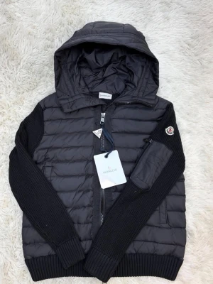 Svart moncler cardigan  - Snygg svart pufferjacka från Moncler med stickade ärmar och huva. Jackan har dragkedja framtill, ficka med dragkedja på ärmen och Moncler-logga på vänster ärm. Materialmix av quiltad nylon och stickad ull ger en modern look. Storlek M men passar oxå för S, Nfc funkar✅