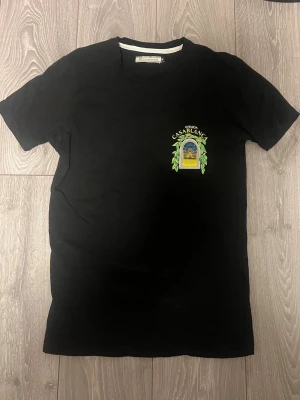 Svart Casablanca t-shirt med tryck - Säljer en svart t-shirt från Casablanca med färgglatt tryck fram och stort grafiskt tryck på ryggen med texten 'Avenida Casablanca' och ett fönster med blad och byggnadsmotiv. Klassisk passform och rund hals. Perfekt för dig som gillar statement-plagg.