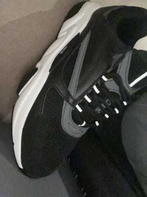 Dior B22 - Snygga svarta sneakers med vit sula och grå reflexdetaljer. Ovandelen är i mesh och syntetmaterial för bra andningsförmåga. Skorna har svarta snören med vita detaljer och en sportig look som passar till streetwear.