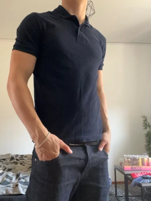 Mörkblå piké t-shirt i bomull - Säljer en stilren mörkblå piké t-shirt med krage och korta ärmar. Tillverkad i mjuk bomull och har en klassisk passform som sitter snyggt på kroppen. Perfekt för dig som gillar en enkel men clean look.