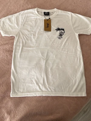 Vit Stüssy dice t-shirt - Vit t-shirt från Stüssy med klassisk logga och två tärningar tryckta på bröstet. T-shirten har rund hals, korta ärmar och är gjord i mjuk bomull. Perfekt för dig som gillar streetwear och vill ha en clean men ändå unik look.