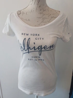 Vit t-shirt från Hilfiger Denim - Vit t-shirt från Hilfiger Denim med tryck 'New York City' och logga framtill. Klassisk rund halsringning och korta ärmar. Liten Hilfiger-flagga på ärmen. Tillverkad i mjuk bomull för en bekväm känsla.