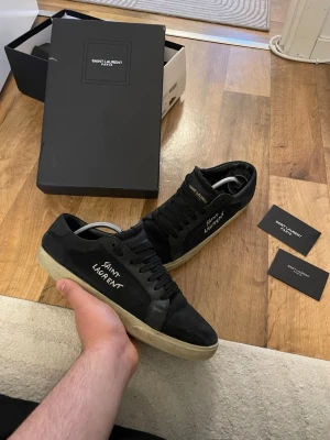 Svarta Saint Laurent sneakers - Svarta sneakers från Saint Laurent med vit logga på sidan och svart mockaöverdel. Skorna har svarta skosnören och en beige sula i gummi. Klassisk låg modell som passar till många olika stilar. Levereras med originalkartong. Obs finns ett litet hål vid hälen  därför pris 