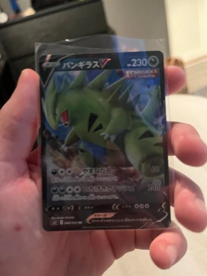 Tyranitar V Pokémonkort japanskt - Japanskt Pokémonkort med Tyranitar V-motiv. Kortet har en cool illustration av den gröna Pokémonen Tyranitar och är inplastat för extra skydd. Texten på kortet är på japanska och det har en mörk bakgrund med blå och röda detaljer.