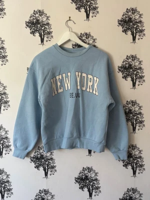 Ljusblå sweatshirt med New York-tryck - Ljusblå sweatshirt med trycket 'NEW YORK EE.UU' i vitt och svart på bröstet. Tröjan har rund halsringning, ribbade muddar vid ärmslut och nederkant samt en avslappnad passform. Perfekt för en casual och trendig look.