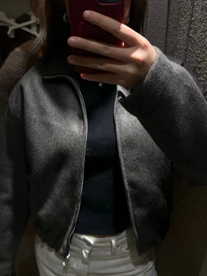 Mörkgrå bomberjacka med dragkedja - Snygg mörkgrå bomberjacka med dragkedja framtill och elastisk midja. Jackan har en klassisk krage och långärmad design, perfekt för en stilren och enkel look. Passar till många olika outfits och är lätt att matcha.