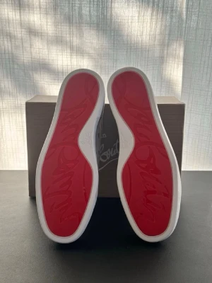 Gråa Christian Louboutin sneakers - Snygga gråa sneakers från Christian Louboutin i mocka med klassiska röda sulor och vita kanter. Skorna har lågt skaft, rund tå och matchande grå skosnören. Perfekta för dig som vill ha en exklusiv och stilren look.